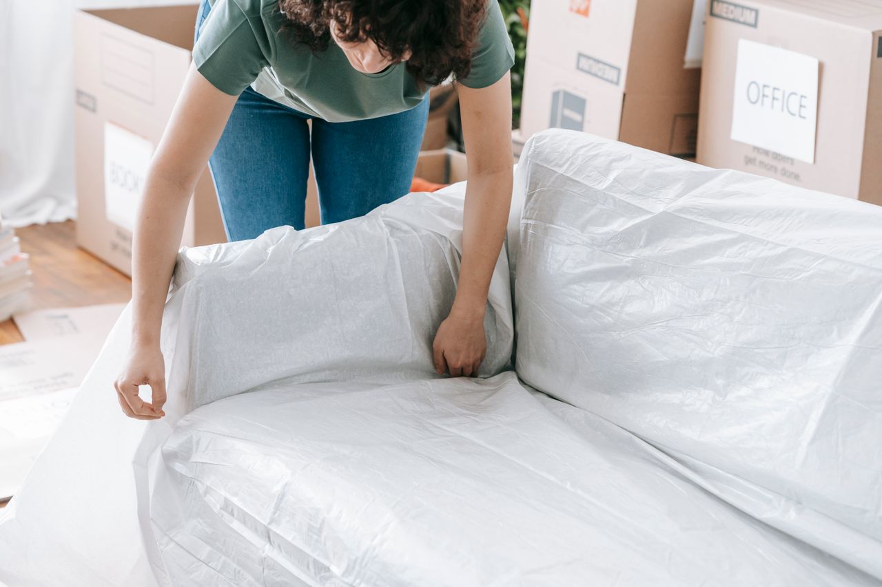 who-we-are A woman carefully wraps a sofa in protective material while packing boxes in a bright room.